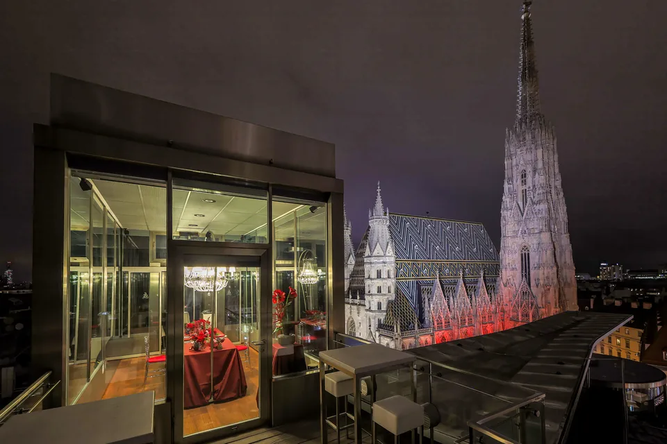 Glaspavillon mit roter Tafel bei Nacht, dahinter der beleuchtete Stephansdom