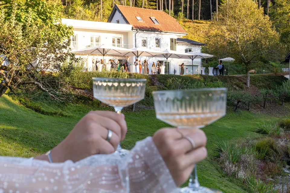 Hochzeitspaar stößt mit Coupé-Gläsern an, dahinter das Haupthaus von Das Haidensee