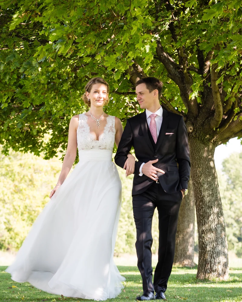 Hochzeitspaar spaziert Arm in Arm unter einem großen Baum im warmen Licht