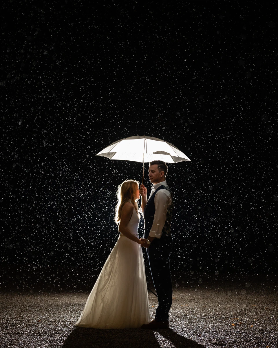 Hochzeitspaar steht bei Nacht im Regen unter einem beleuchteten Schirm