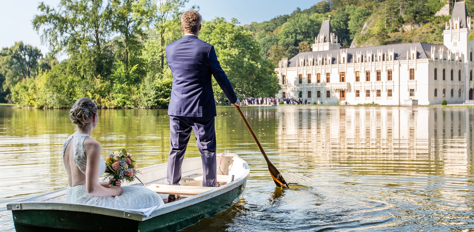 Hochzeitspaar im Ruderboot am Teich vor Schloss Hernstein, die Gäste warten am Ufer