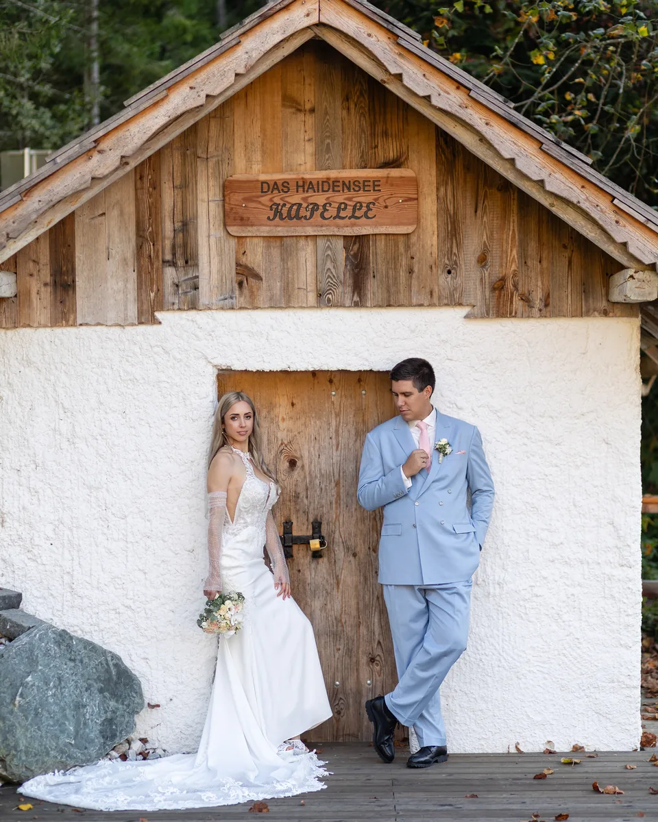 Hochzeitspaar lehnt an der weißen Holzkapelle am Haidensee, über der Tür das Schild der Kapelle