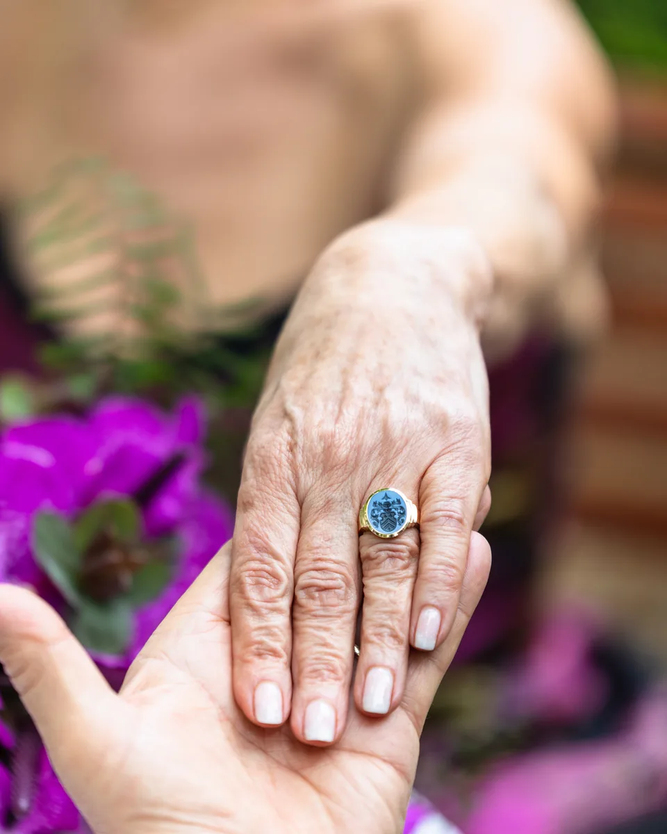 Hand mit einem Ring mit blauem Stein in der Hand des Partners, dahinter violette Blumen
