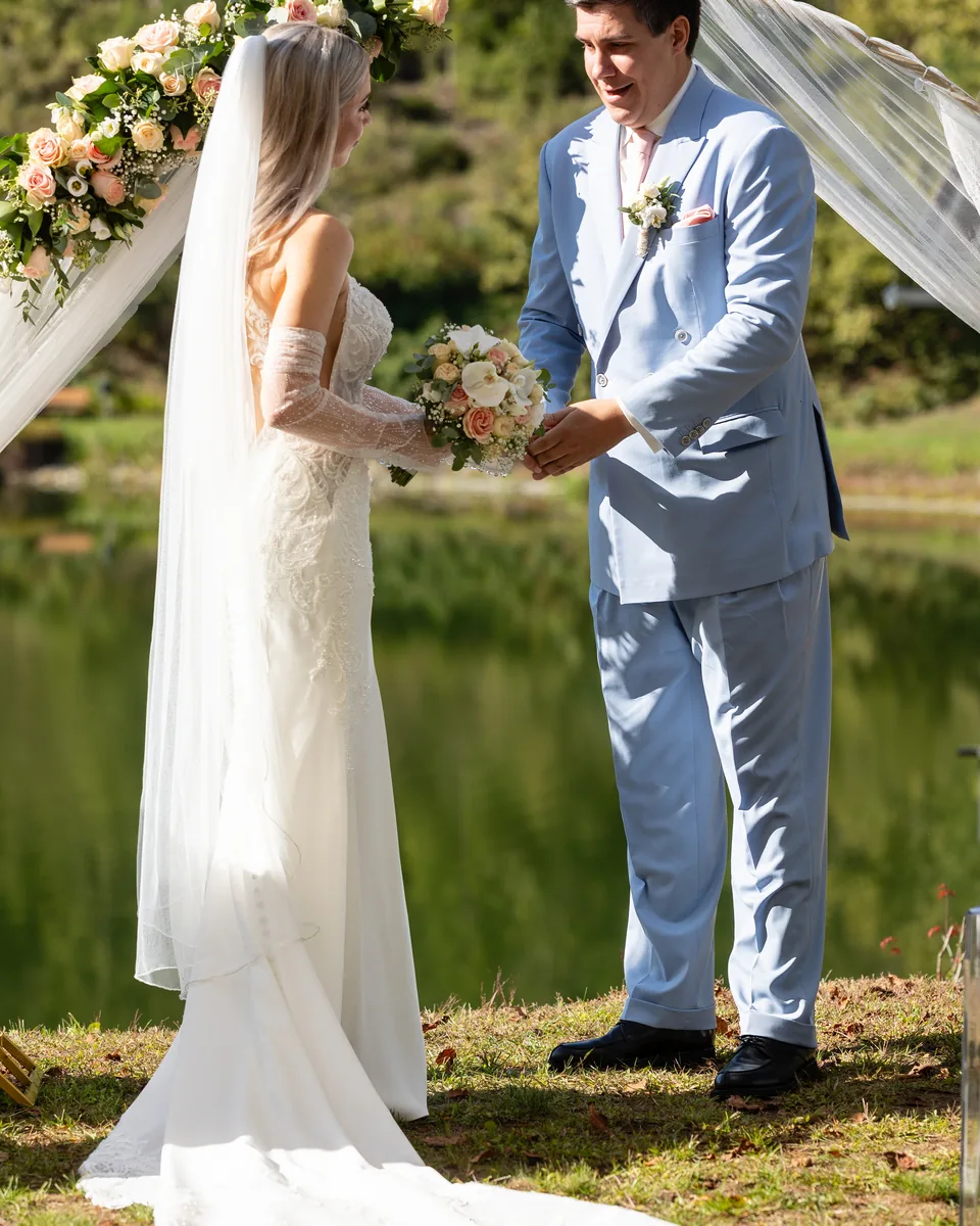 Hochzeitspaar hält sich beim Eheversprechen an den Händen, hinter ihnen der Blumenbogen und der See