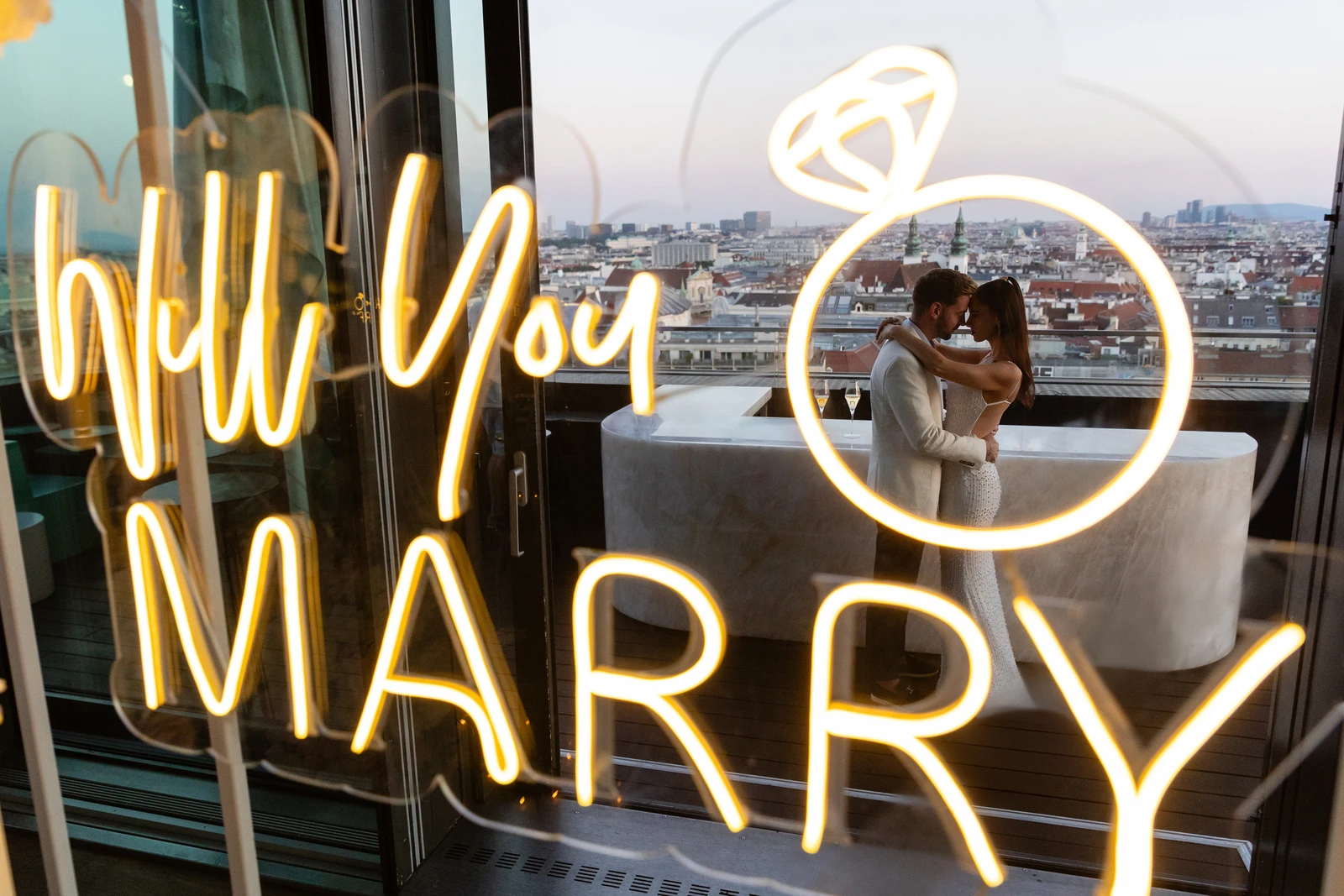 Will You Marry Me neon sign, with the newly engaged couple above the rooftops of Vienna