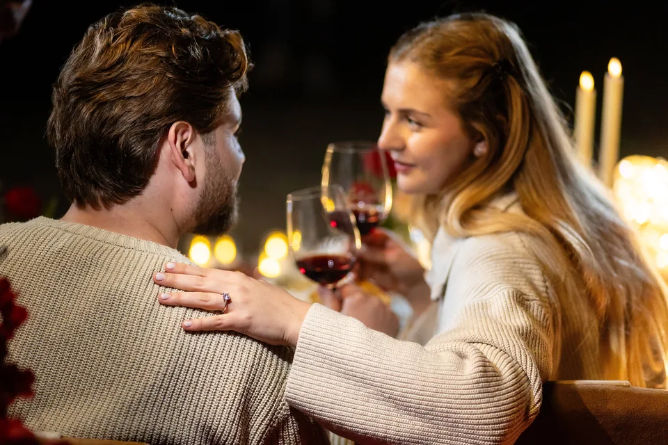 Frisch verlobtes Paar stößt mit Rotwein an, ihre Hand mit Ring auf seiner Schulter