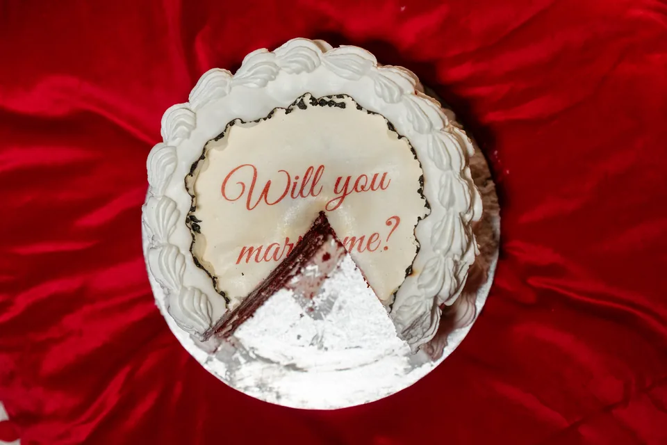 Torte mit der Aufschrift Will you marry me auf rotem Samtkissen, darunter die Verlobungsringe