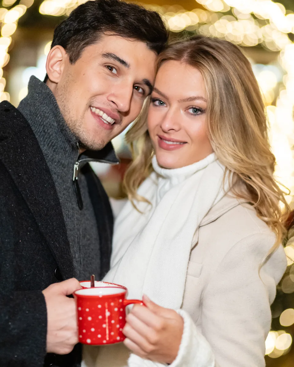 Paar mit roten Punschhäferln auf einem Wiener Weihnachtsmarkt, dahinter Lichterketten